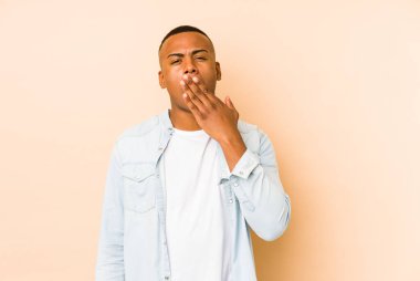 Genç latin adam bej arka planda izole edilmiş esniyor. Ağzı eliyle kapatan yorgun bir hareket sergiliyor..