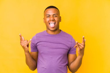 Sarı arka planda izole edilmiş genç latin adam parmaklarıyla kaya hareketi gösteriyor.