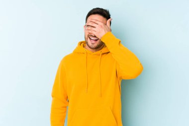 Mavi arka planda soyutlanmış genç beyaz bir adam elleriyle gözlerini kapatıyor, geniş bir gülümsemeyle bir sürpriz bekliyor..