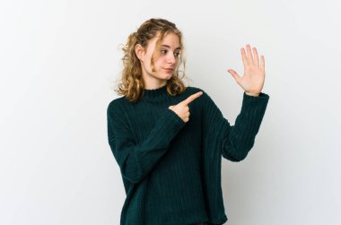 Beyaz arka plandaki genç beyaz kadın gülümseyerek beş numarayı parmaklarıyla gösteriyor..