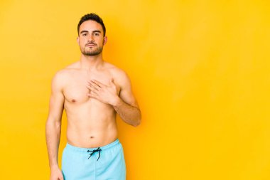 Sarı arka planda mayosu olan genç beyaz bir adam. Yeminli beyaz bir genç, elini göğsüne koyuyor..