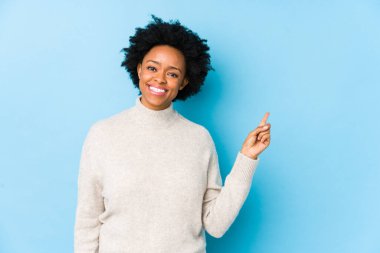 Orta yaşlı Afro-Amerikalı bir kadın, mavi arka planda yalnız ve mutlu bir şekilde işaret parmağıyla işaret ediyor..