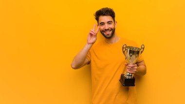 Genç adam zafer jesti yaparak elinde bir kupa tutuyor.