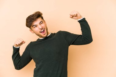 Genç Arap adam bej bir arka planda izole edilmiş zafer kazandıktan sonra yumruğunu kaldırıyor, kazanan konsept..