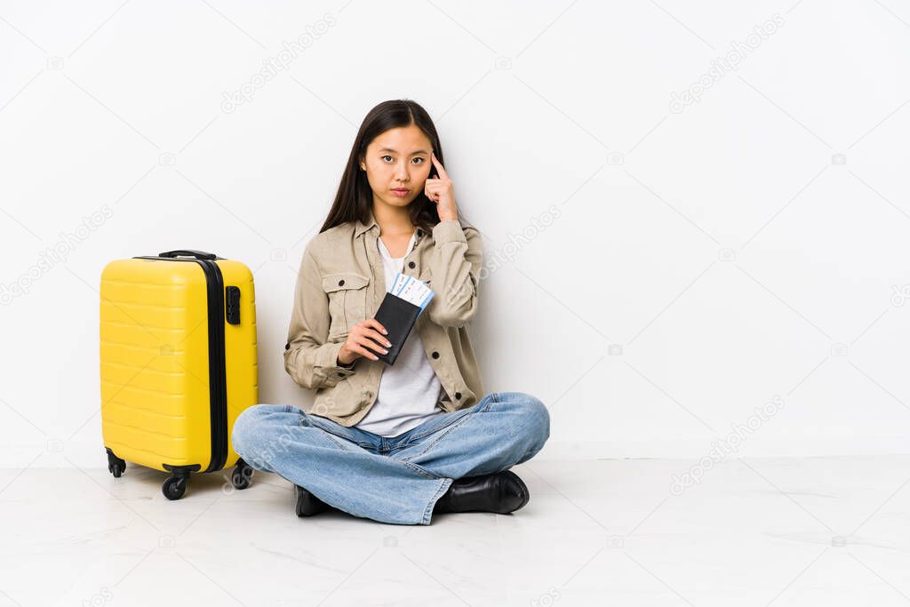 Young chinese traveler woman sitting holding a boarding passes pointing temple with finger, thinking, focused on a task.