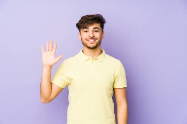 Mor arka planda izole edilmiş genç bir Arap adam gülümseyerek beş numarayı parmaklarıyla gösteriyor..