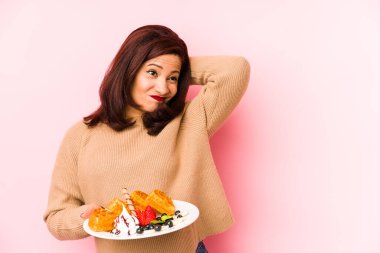 Orta yaşlı, elinde waffle tutan Latin bir kadın. Kafasının arkasına dokunuyor, düşünüyor ve bir seçim yapıyor..