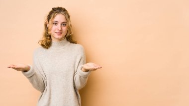 Beyaz tenli genç bir kadın bej arka planda izole edilmiş, kollarıyla ölçek yapıyor, mutlu ve kendinden emin hissediyor..