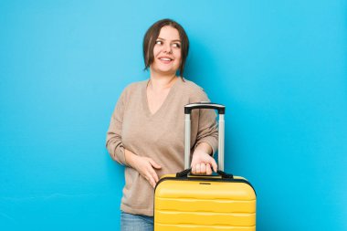 Elinde bavul tutan kıvrımlı bir kadın gülümseyen, neşeli ve hoş bir şekilde yana bakıyor..