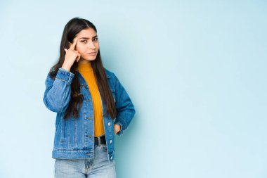 Genç Hintli bir kadın mavi arka planda izole edilmiş. Parmağıyla tapınağı işaret ediyor. Düşünüyor, bir göreve odaklanıyor..