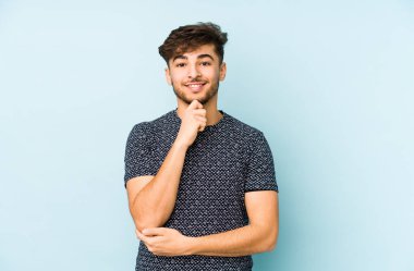 Mavi bir arka planda izole edilmiş genç bir Arap adam gülümsüyor ve kendinden emin, elleriyle çenesine dokunuyor..