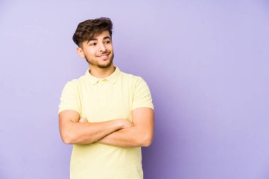 Mor arka planda soyutlanmış genç bir Arap adam kollarını kavuşturarak gülümsüyor..