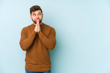 Kafkasyalı genç adam mavi arka planda izole edilmiş dua ediyor, bağlılık gösteriyor, dindar biri ilahi ilham arıyor..