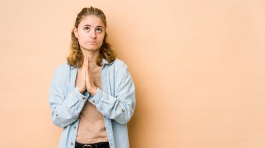 Beyaz tenli genç bir kadın, bej arka planda izole edilmiş, el ele tutuşmuş, ağız kenarında dua ediyor, kendinden emin..