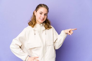 Mor arka planda genç beyaz kadın işaret parmağıyla işaret ederek gülümsüyor..