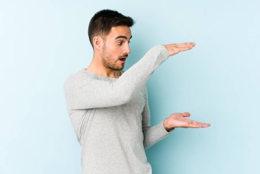 Mavi arka planda izole genç beyaz adam şok ve hayret içinde elleri arasında bir kopya boşluk tutuyor.
