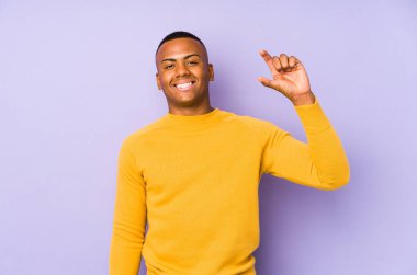 Mor arka planda izole edilmiş genç bir latin adam işaret parmaklarıyla küçük bir şey tutuyor, gülümsüyor ve kendinden emin..