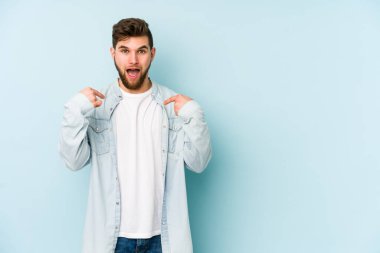 Mavi arka planda izole edilmiş genç beyaz adam parmağını göstererek şaşırdı, geniş bir gülümsemeyle..
