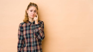 Beyaz tenli genç bir kadın bej arka planda izole edilmiş ağlıyordu, bir şeyden mutsuzdu, acı ve kafa karışıklığı içeriyordu..