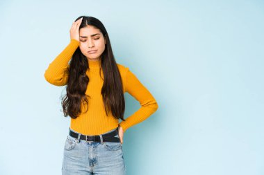 Mavi arka planda izole edilmiş genç Hintli kadın yorgun ve uykulu ellerini başının üstünde tutuyor..