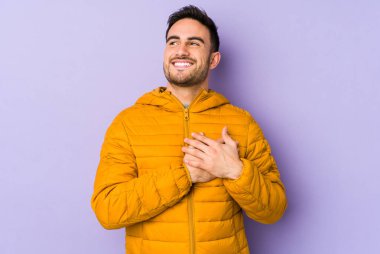 Mor arka planda izole olmuş genç beyaz adam gülüyor, ellerini yüreğine koyuyor, mutluluk kavramını koruyor..