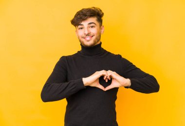 Sarı arka planda izole edilmiş genç bir Arap adam gülümsüyor ve elleriyle kalp şeklini gösteriyor..
