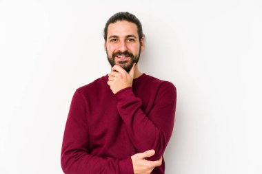 Uzun saçlı genç adam beyaz bir arka planda izole edilmiş mutlu ve kendinden emin gülümsüyor, elleriyle çenesine dokunuyor..