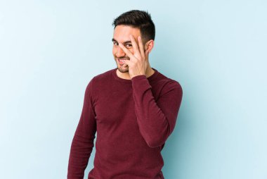 Mavi arka planda izole edilmiş genç beyaz adam kameraya parmaklarıyla göz kırpıyor, yüzünü kapatmaktan utanıyor..