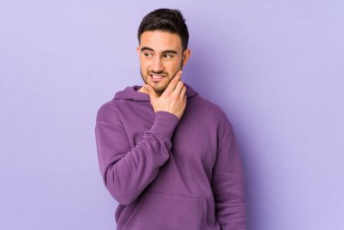 Kafkasyalı genç bir adam mor arka planda izole edilmiş kafasının arkasına dokunuyor, düşünüyor ve bir seçim yapıyor..