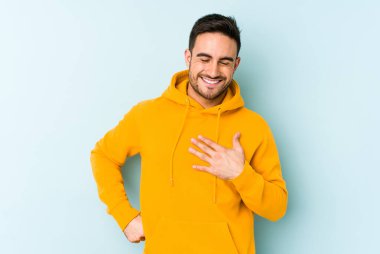 Kafkasyalı genç adam mavi arka planda izole olmuş gülüyor, elini yüreğine koyuyor, mutluluk kavramını koruyor..