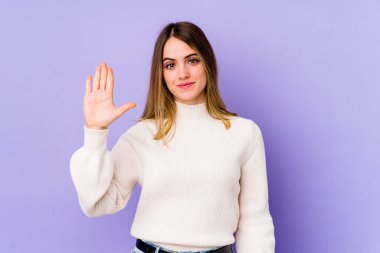 Mor arka planda izole edilmiş genç beyaz kadın gülümseyerek beş numarayı parmaklarıyla gösteriyor..