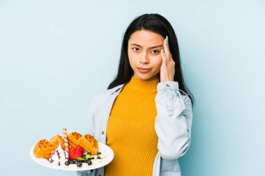 Mavi arka planda waffle tutan genç Çinli kadın işaret parmağıyla yaptığı hayal kırıklığı jestini gösteriyor..