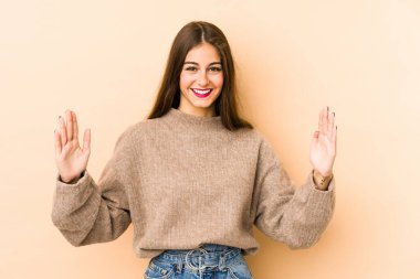 Beyaz tenli genç bir kadın bej arka planda izole edilmiş. Elinde parmakları olan, gülümseyen ve kendinden emin bir şey var..