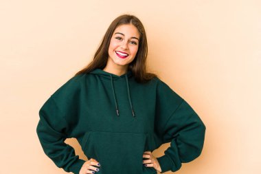 Beyaz tenli genç bir kadın, bej arka planda izole edilmiş. Kendine güvenen, ellerini kalçasından ayırmayan..