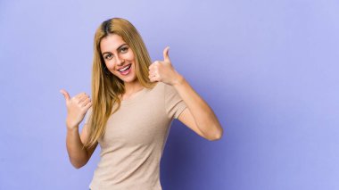 Mor arka planda izole edilmiş genç sarışın kadın iki parmağını da kaldırıyor, gülümsüyor ve kendinden emin..