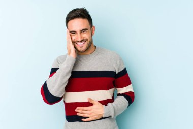 Mavi arka planda izole edilmiş genç beyaz adam mutlu bir şekilde gülüyor ve ellerini karnının üstünde tutarak eğleniyor..