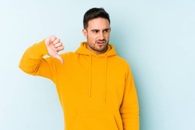 Mavi arka planda izole edilmiş genç beyaz bir adam baş parmağını aşağı gösteriyor ve hoşlanmadığını ifade ediyor..