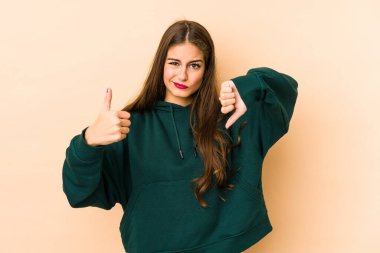 Beyaz tenli genç bir kadın bej arka planda izole edilmiş. Baş parmaklarını kaldırmış, baş parmağını aşağı gösteriyor. Zor seçim konsepti.
