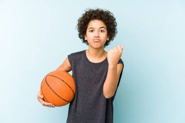 Basketbol oynayan çocuk mavi arka planda izole edilmiş kameraya yumruk gösteriyor, agresif yüz ifadesi var..