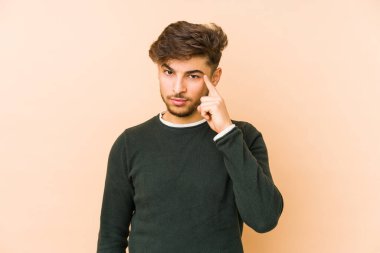 Bej bir arka planda izole edilmiş genç bir Arap adam şakağı parmağıyla işaret ediyor, düşünüyor ve bir göreve odaklanıyor..