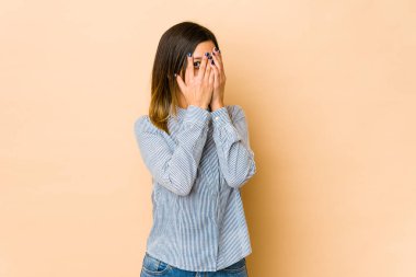 Bej arka planda izole edilmiş genç bir kadın parmaklarının arasından korkmuş ve gergin bir şekilde göz kırpıyor..