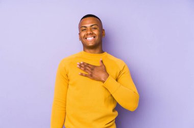 Mor arka planda izole edilmiş genç latin adam yüksek sesle gülüyor elini göğsüne koyuyor.