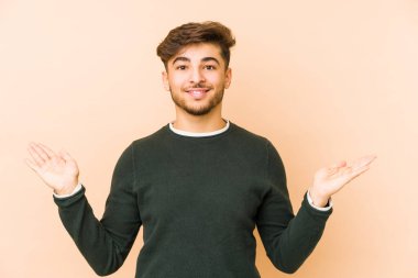 Bej bir arka planda izole genç bir Arap adam kollarıyla ölçek yapar, mutlu ve kendinden emin hisseder..