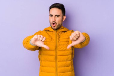 Mor arka planda izole edilmiş genç beyaz adam baş parmağını aşağı gösteriyor ve hoşlanmadığını ifade ediyor..