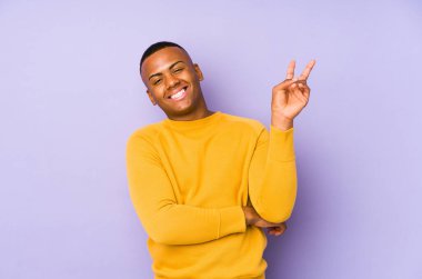 Mor arka planda izole edilmiş genç Latin adam neşeli ve kaygısız parmakları olan bir barış sembolü gösteriyor..