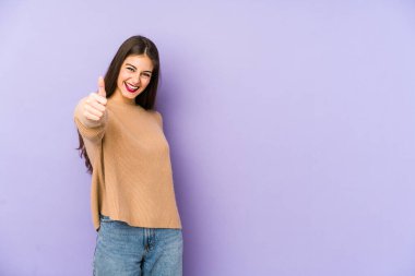 Mor arka planda izole edilmiş genç beyaz kadın gülümsüyor ve başparmağını kaldırıyor.