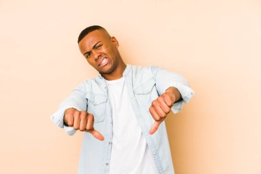 Bej arka planda izole edilmiş genç latin adam baş parmağını aşağı gösteriyor ve hoşlanmadığını ifade ediyor..