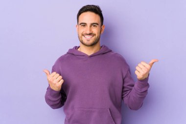 Mor arka planda izole edilmiş genç beyaz adam iki parmağını da kaldırıyor, gülümsüyor ve kendinden emin..