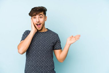 Mavi arka planda izole edilmiş genç bir Arap, avuç içinde kopya alanı tutuyor, el üstünde tutuyor. Şaşırmış ve mutlu..