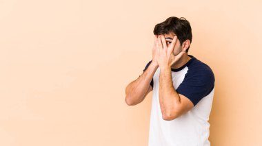 Bej arka planda izole edilmiş genç adam parmaklarının arasından korkmuş ve gergin bir şekilde göz kırp..